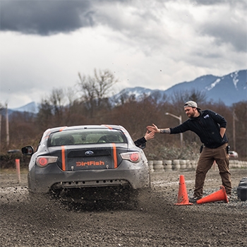 View of the back end of a Subaru on a gravel course moving past some orange cones. On the passenger side, a hand is reaching out from the window and high-fiving the hand of a man standing behind the cones. Where the license plate would usually be placed, it says DirtFish in orange.