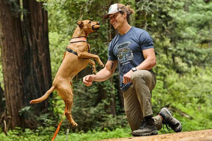 A dog is jumping up and catching a toy in its mouth. A male, who is wearing a Wild Tribute cap and tee, is kneeling in front of the dog and reaching out to it, smiling.