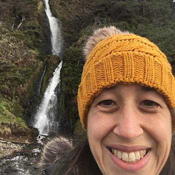 A selfie of Katie Buckles, smiling and wearing a knit hat with a tassel. Behind her, a waterfall cascades down a mossy, green mountainside. 