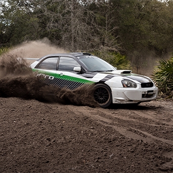 A racing car with RallyPro on the side is driving at a high speed through gravel, kicking up a thick cloud of black dust.