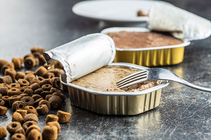 Two cans of pet food on a table with loose kibble behind them. A fork is resting on the first open can of food.