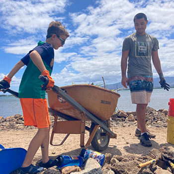 Nikko is pushing a wheelbarrow on a sandy path near the ocean’s edge. A Hawaiian resident, who appears to also be working, is standing nearby. 