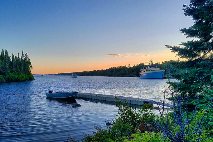 A large blue boat is in the far right of the frame with a pinkish sunset behind it. In the foreground is a dock with a small boat with trees and shrubs on both sides of the frame.