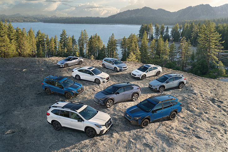 In a wilderness setting, the 2023 Subaru family of vehicles are parked in a circle on a land clearing. In the background are evergreen trees, a body of water and mountains.