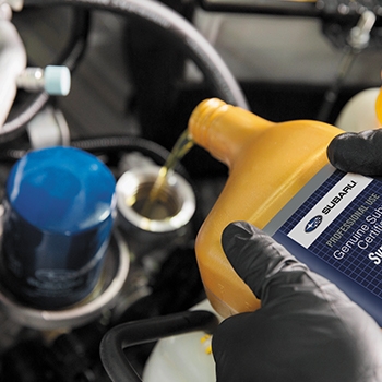 Two hands are holding a fresh bottle of Subaru Synthetic Motor Oil and pouring the oil into a Subaru vehicle.