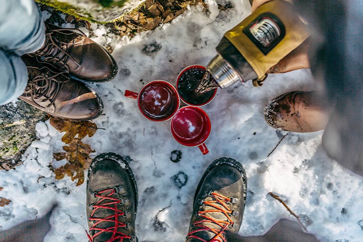 Closeup of the boots of three people who are standing in snow. One person is pouring a hot refreshment into one of three red cups in the center.