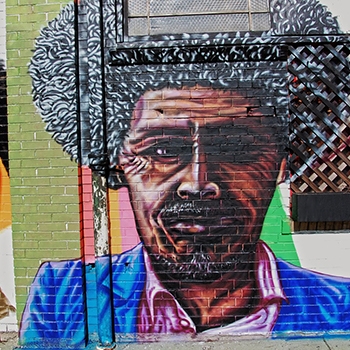 Artwork on the side of the building of a middle-aged Black man wearing a blue jacket with a lapel and a pink shirt with a collar.
