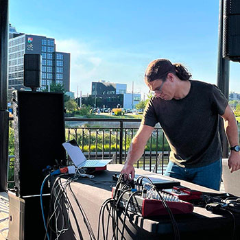 Chris Heier performing original techno music live at the East Village Gazebo in Calgary, Alberta, Canada. Modern buildings can be seen in the distance beyond the gazebo’s sleek metal railings.