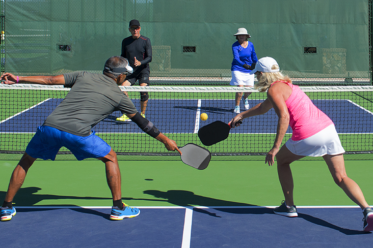 Two couples are actively playing a game of pickleball as doubles. They're dressed for warm weather in shorts and brimmed hats or visors.