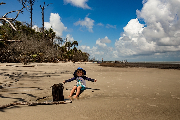 The Jacobson’s child sitting in the sand near the shoreline wearing a hat, shorts and a long-sleeved shirt during their stay on Little Tybee Island, Georgia.