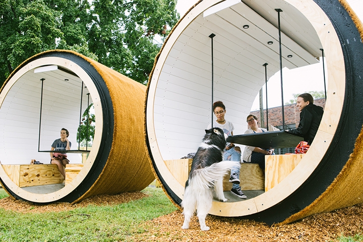 A black and white dog jumps up to a group of people at a picnic-style table set into a large cylinder at the dog park.