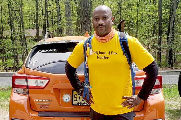 Kasim Carter is standing in front of his orange Subaru Crosstrek. He has his hands resting on his hips and is wearing a yellow T-shirt that says Outdoor Afro Leader and a backpack. There are deciduous trees in the distance.
