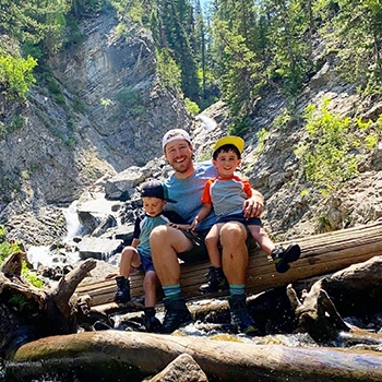 A smiling man and his sons sitting on a log with a waterfall behind them.