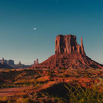 A large red landform, called a butte, that is known as Monument Valley, Arizona, stands out in the distance with a gorgeous blue sky behind it.