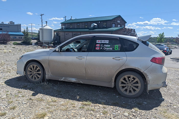 A muddy 2008 Subaru WRX sits in a gravel parking lot in front of the Half Hitch Brewing Company building.