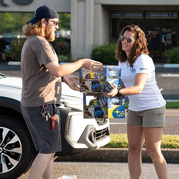 Lindsay Forney is shown working on behalf of Cruise for Foose. She appears to be collecting two boxes of toy Subaru cars from a male. They're both wearing shorts and T-shirts.