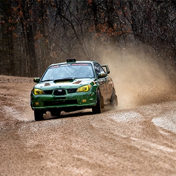 A green vehicle is driving on a muddy gravel road. Behind it, a trail of mud and gravel is being kicked up by the wheels.