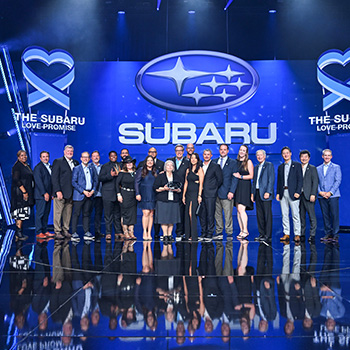 About 20 representatives from Subaru of Las Vegas are standing on a stage with the Subaru Love Promise and Subaru logo behind them accepting the 2023 Subaru Love Promise Retailer of the Year Award. They are wearing dressy business attire.