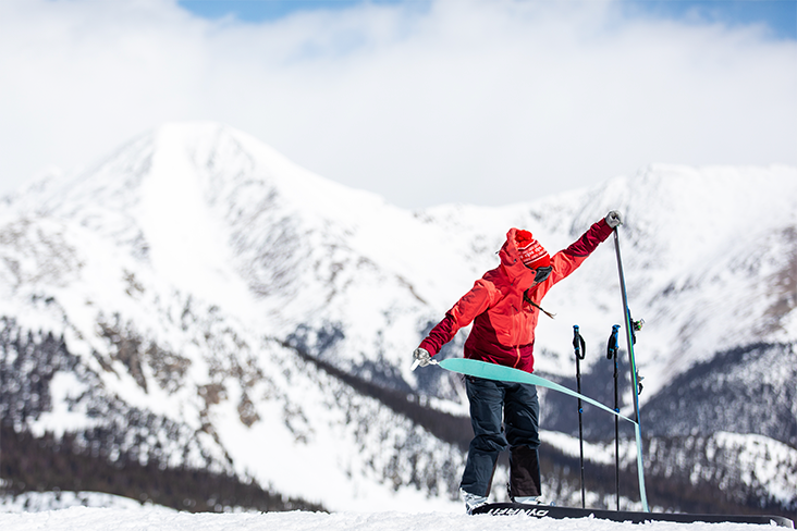 A person is making an adjustment on their ski. There are snow-capped mountains in the distance.