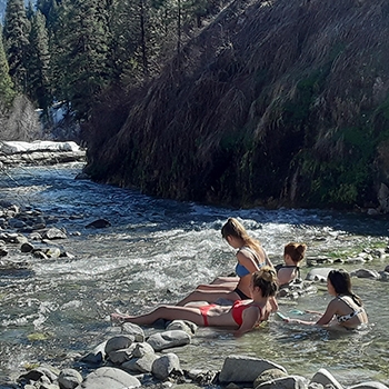 The four teenagers, wearing swimsuits, are relaxing in a hot spring. Rocky cliffs are about 10 feet to the right and a cluster of evergreen trees are farther off in the distance.