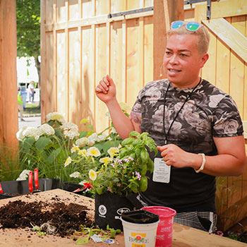 Botanical artist Tu Bloom is standing behind a large wooden table that is filled with loose dirt, pots and various plant and flower varieties. He is holding up one hand and appears to be speaking to attendees of a Subaru Potting Party.