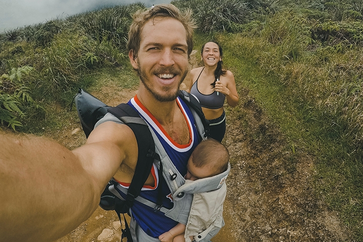 Man holding his infant in a front carrier while on a hike with a woman, smiling.