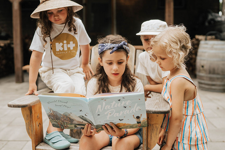 Several children are gathered around a book, titled Nature Parade, with the oldest child in the center reading from it.