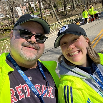 Mark Grady is standing on the street during the Boston Marathon. He's with his water stop co-captain, Courtney Turner Hill. They're both smiling and wearing reflective coats in bright yellow.