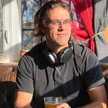 Chris Heier, a long-haired brunette Caucasian male in wire-rimmed glasses and a gray short-sleeved shirt, is enjoying a beverage on a brown leather couch in the sunshine. Behind him, a window with the curtains pulled back can be seen, and the sunshine appears to be coming from a window out of view to the right.