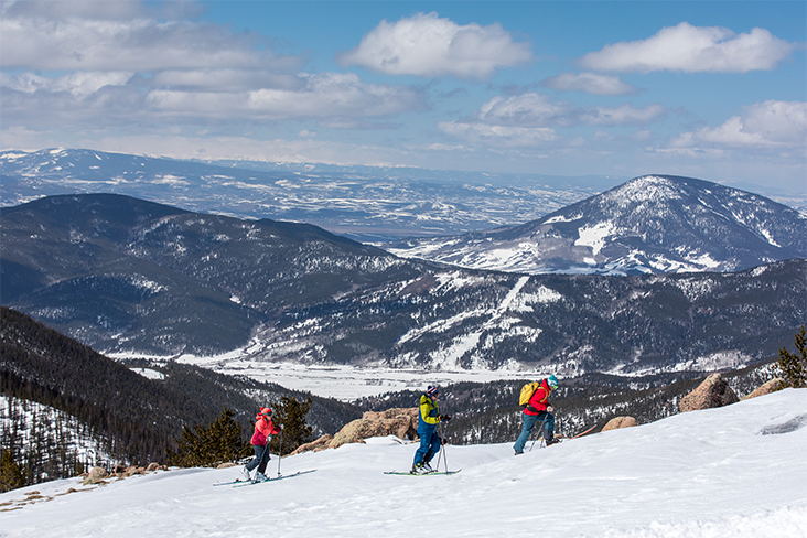 A view of three people uphilling in thick snow. In the distance, mountains partially covered in snow dominate the landscape.