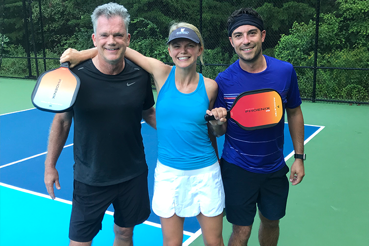 Hannah and Tobi Lutz, holding pickleball rackets, are on a pickleball court with another pickleball player. They are all smiling and joined arm in arm.