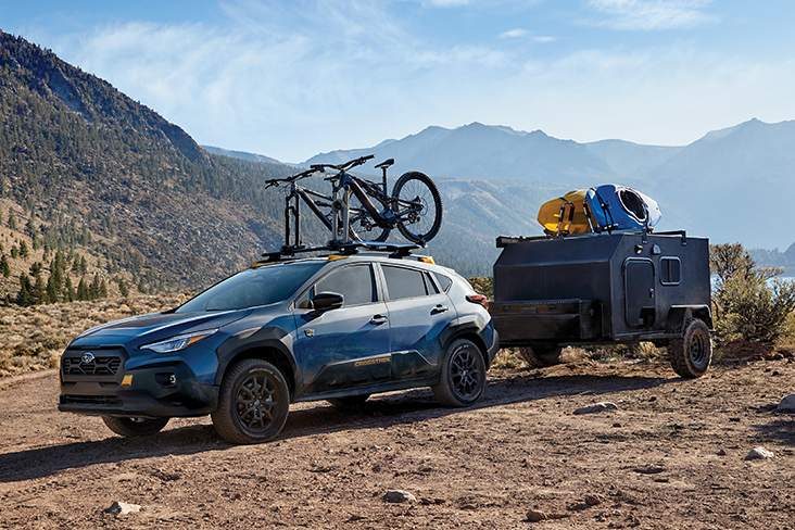 A Subaru Crosstrek Wilderness on a dirt road with mountains in the background. The Crosstrek Wilderness has two bicycles on the rooftop and is towing a small trailer with two kayaks on the top.