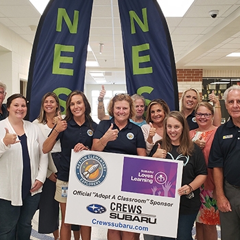 About 10 are people standing around a sign that says, “Official Adopt A Classroom Sponsor, Crews Subaru, crewssubaru.com.” Everyone is holding a thumb up to signify their approval; they look happy and are smiling.