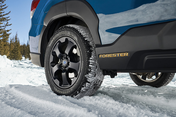 Close-up of the back tires of a Subaru Forester Wilderness in the snow. Evergreen trees can be seen in the distance.