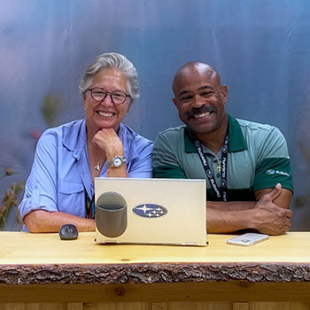 Denise Coogan, Environmental Partnership Manager, and Anthony Trosclair, Brand Partnerships Specialist, are seated at a wooden table, smiling. In front of them on the table is a Subaru laptop with a mouse next to it.
