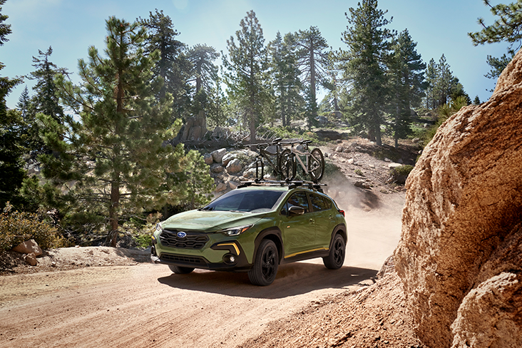 A 2024 Subaru Crosstrek Sport is climbing a rough dirt trail with a large boulder on the right and evergreens and loose rocks on the left. The Crosstrek Sport features a bike rack on top with two bicycles.