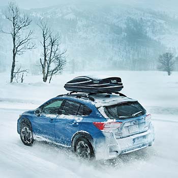 A 2022 Subaru Crosstrek in Horizon Blue with a Thule Cargo Carrier secured to the rooftop is kicking up snow on a snowy road with snow-covered mountains in the distance.