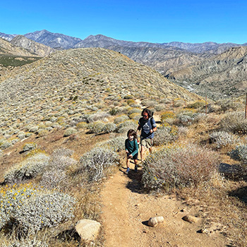 Jason Yasuda and son, Everest Yasuda, hiking a segment of the Pacific Crest Trail in Whitewater, California. The dirt trail winds through desert shrubs, and mountains can be seen in the distance.