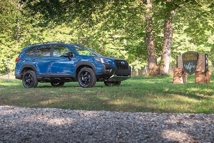 The 2022 Forester Wilderness parked on grass at the Subaru Wilderness campsite with pebble gravel path in front of the vehicle and tall evergreens behind it.
