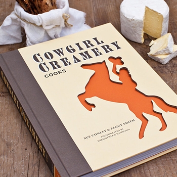 A Cowgirl Creamery cookbook with an illustration on the front of a cowgirl waving a hat and riding a bucking horse.