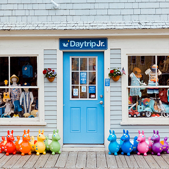 An outdoor view of the front door to Daytrip Jr. The door is a sky-blue color, and a line of colorful toys can be seen on each side of the door under large windows with children's clothing displayed.