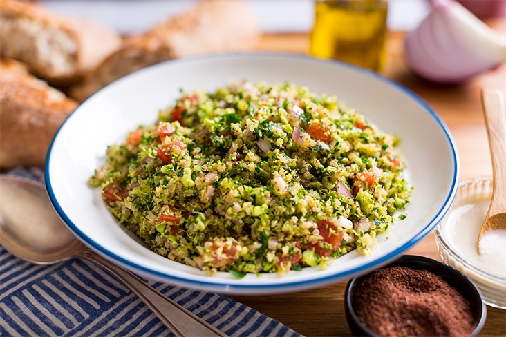 White ceramic bowl with a blue rim filled with the broccoli tabbouleh. The bowl is resting on a wooden table with small loaves of bread in the background and a small dish with ground cumin in the foreground.
