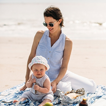 Hailey, a white woman with brown hair, sits on a blanket on the beach, looking down at baby Land, 7 months old, wearing a gray onesie and a white patterned hat. Hailey wears a sleeveless chambray button down shirt, white pants and sunglasses.