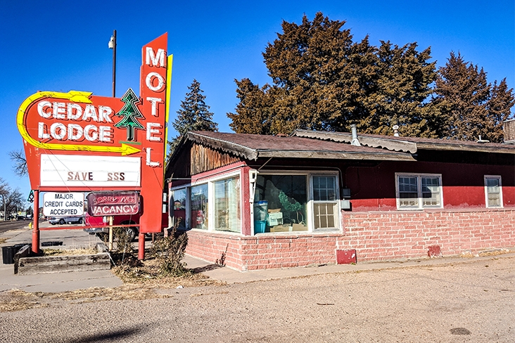 A single-story motel in red brick with a weathered sign on the sidewalk in front of it that says, Cedar Lodge Motel and Major Credit Cards Accepted. The upper halves of large deciduous trees can be seen past the roof of the motel.