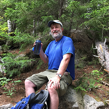 Smith is resting on a large boulder with a bottle of water in one hand. He is wearing a cap, shorts and T-shirt.