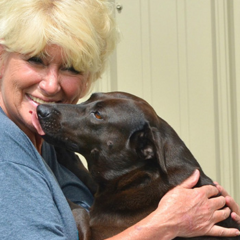Subaru owner Sandy Williams, who has short blonde hair and is wearing a blue shirt, smiles as a black Lab licks her face. 