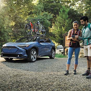 A couple looks over a map while standing near their Subaru Solterra on a gravel road with mature trees in the background.