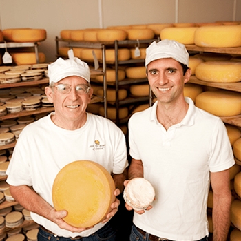 Two cheesemakers, each holding a cheese wheel. In the background are cheese wheels stacked on several carts.