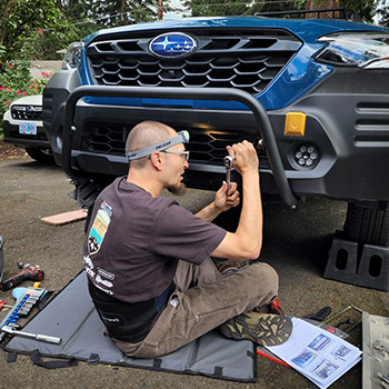 Andy Lilienthal is sitting on a pad in front of the Subaru Outback Wilderness in a driveway. He is installing a Rally Innovations light bar to the vehicle using a socket wrench.