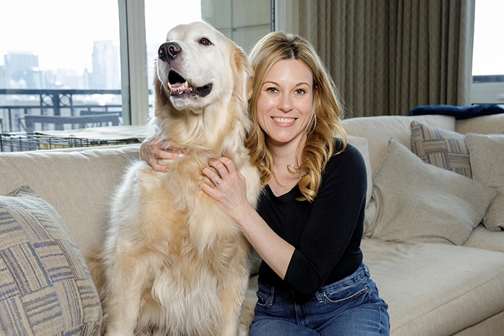 Erin sitting with her gold retriever, Gatsby, on a couch, with high-rise window views of a city.